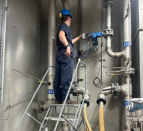 A worker in safety gear stands on a metal step ladder and adjusts a valve on industrial pipes inside a facility.