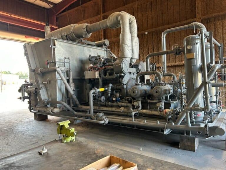 Large industrial compressor unit with multiple pipes, valves, and insulation, situated inside a warehouse with a partially open door.