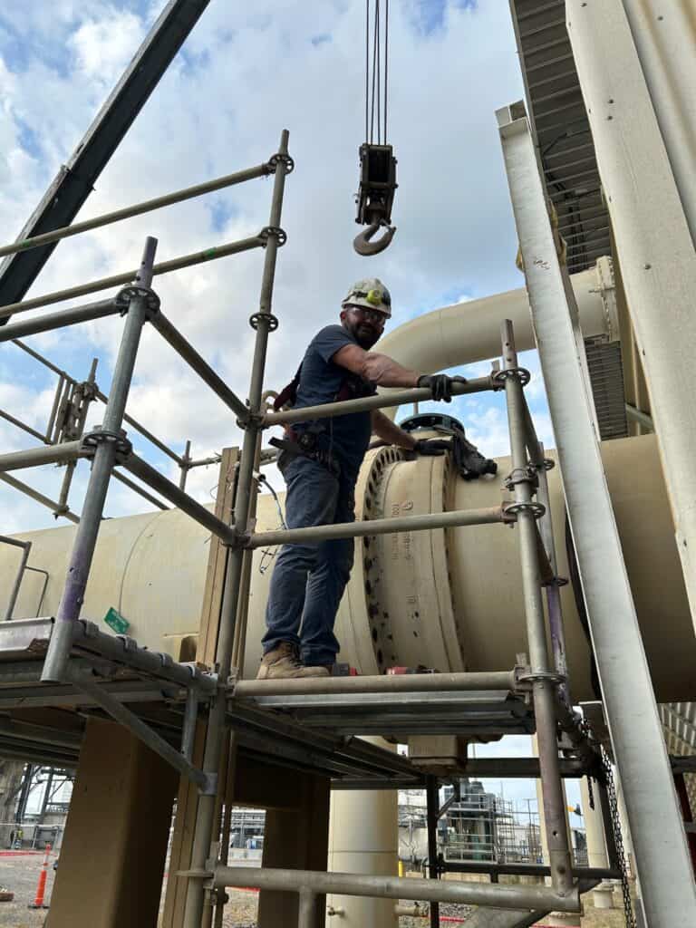 A construction worker wearing safety gear stands on scaffolding near large industrial pipes, holding onto a metal bar.