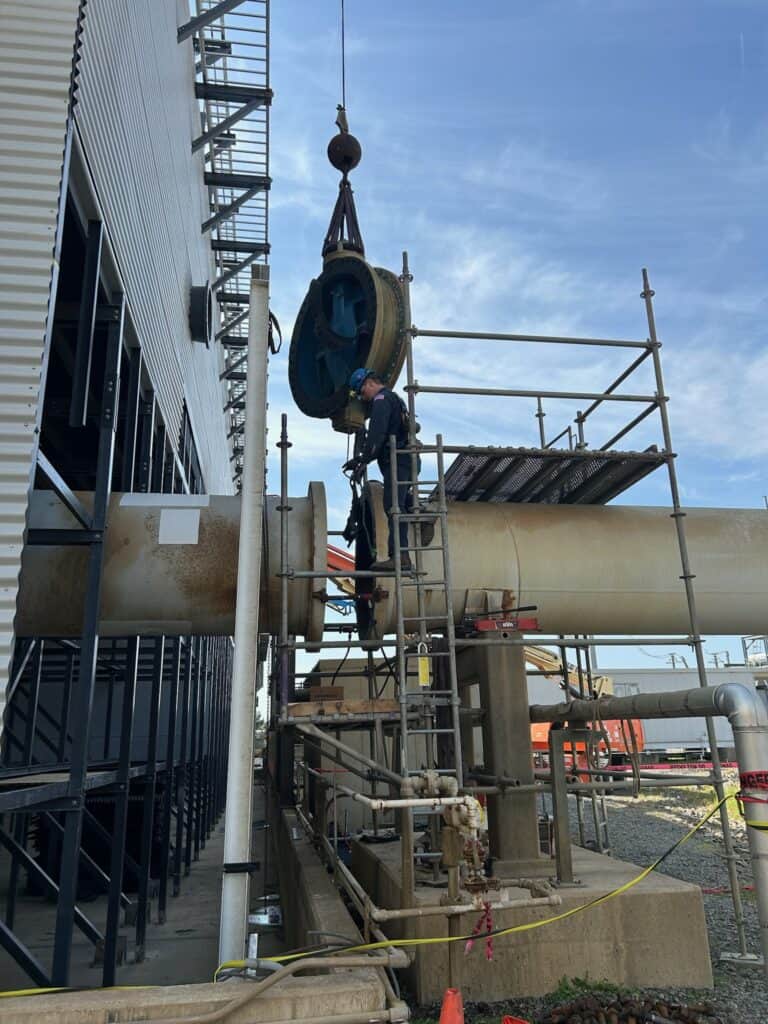 A worker stands on scaffolding, guiding a large pipe flange being lifted by a crane at an industrial construction site.