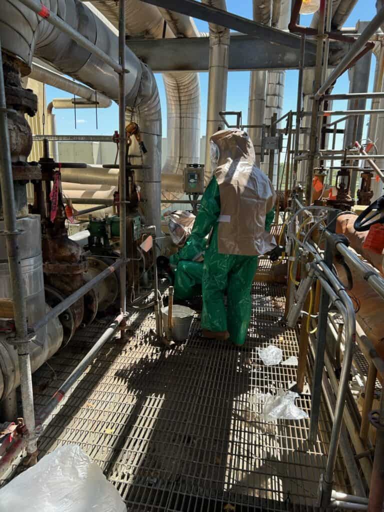 Two people wearing green hazmat suits work on industrial equipment surrounded by pipes and scaffolding in an outdoor setting.