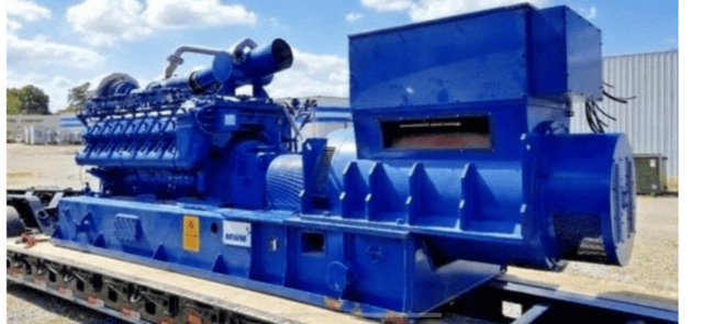A large industrial blue machine with multiple pipes and vents is mounted on a flatbed trailer outdoors.