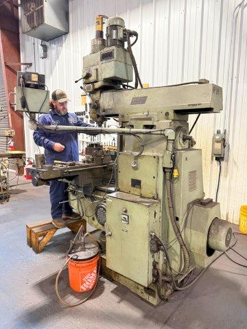 A man operates a large industrial milling machine in a workshop with metal walls and concrete floor.