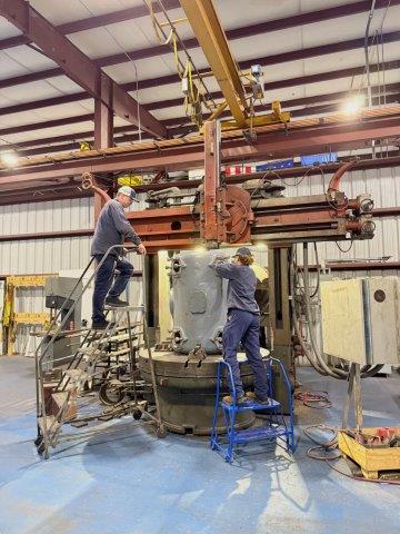 Two workers in safety gear stand on ladders operating machinery around a large cylindrical metal part in an industrial workshop.