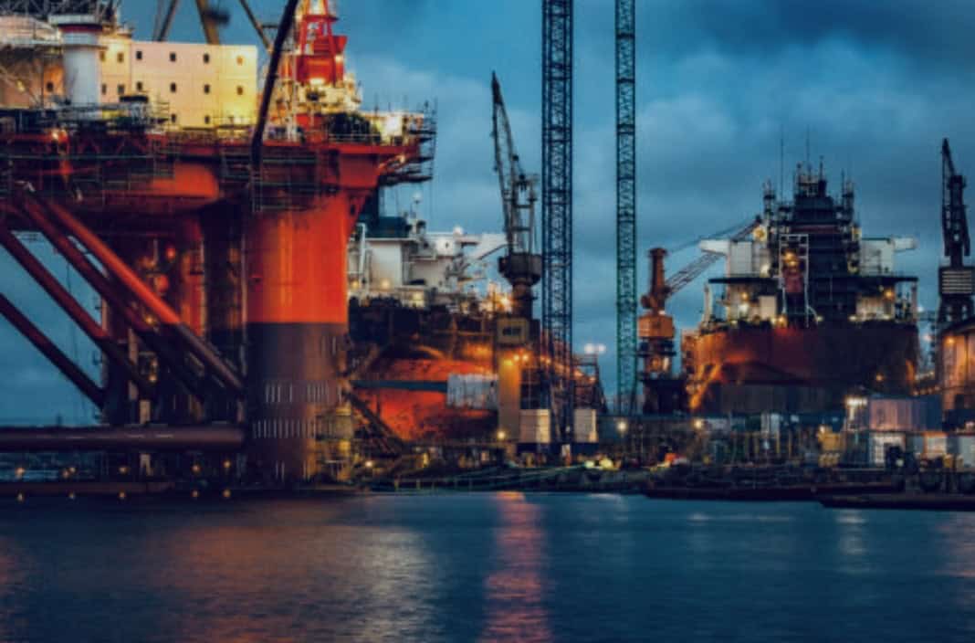 Large offshore oil rigs and cranes are illuminated at dusk, with water in the foreground under a cloudy sky.