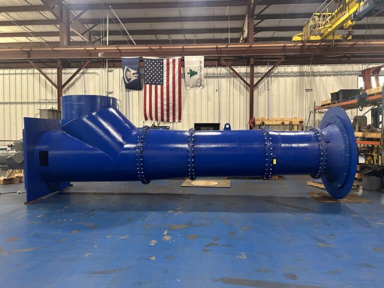 A large, blue industrial pipe with flanges is positioned horizontally inside a warehouse with flags hanging on the back wall.