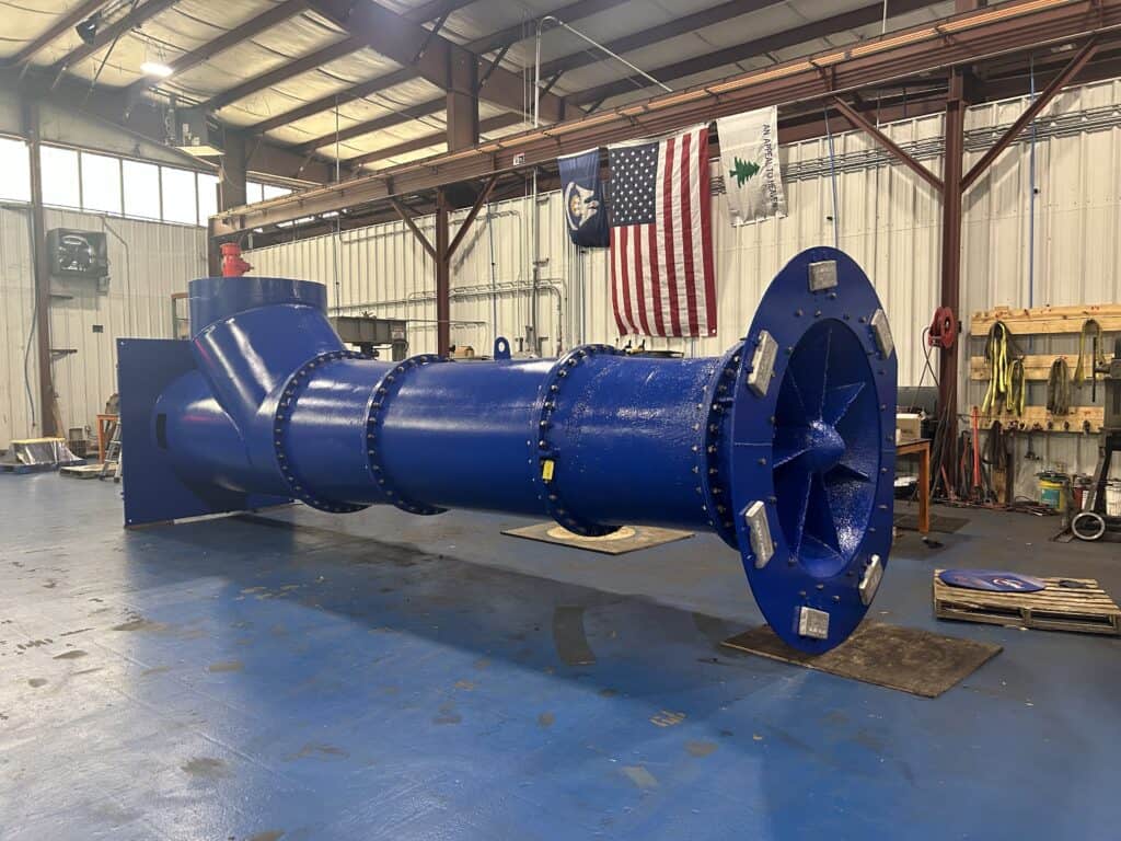 Large blue industrial machine part, possibly a pump or turbine, inside a warehouse with flags and equipment in the background.