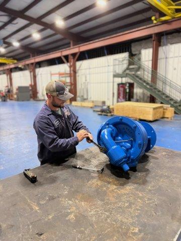 A man in a workshop inspects or assembles a large blue industrial pump on a workbench.