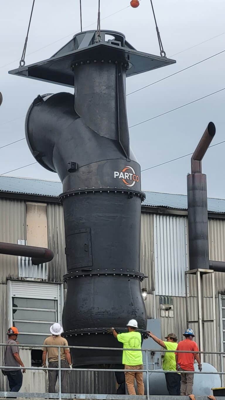 Five workers guide a large black industrial pipe into place outside a metal building using a crane.