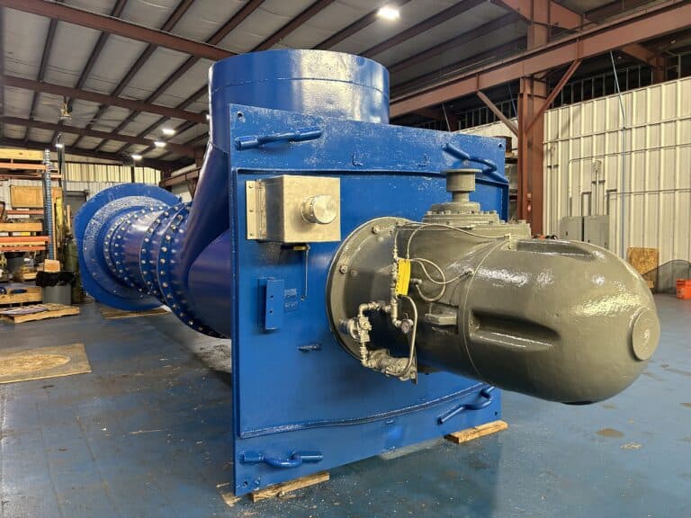 Large industrial hydraulic valve with blue and gray metal components inside a warehouse with shelves and tools in the background.