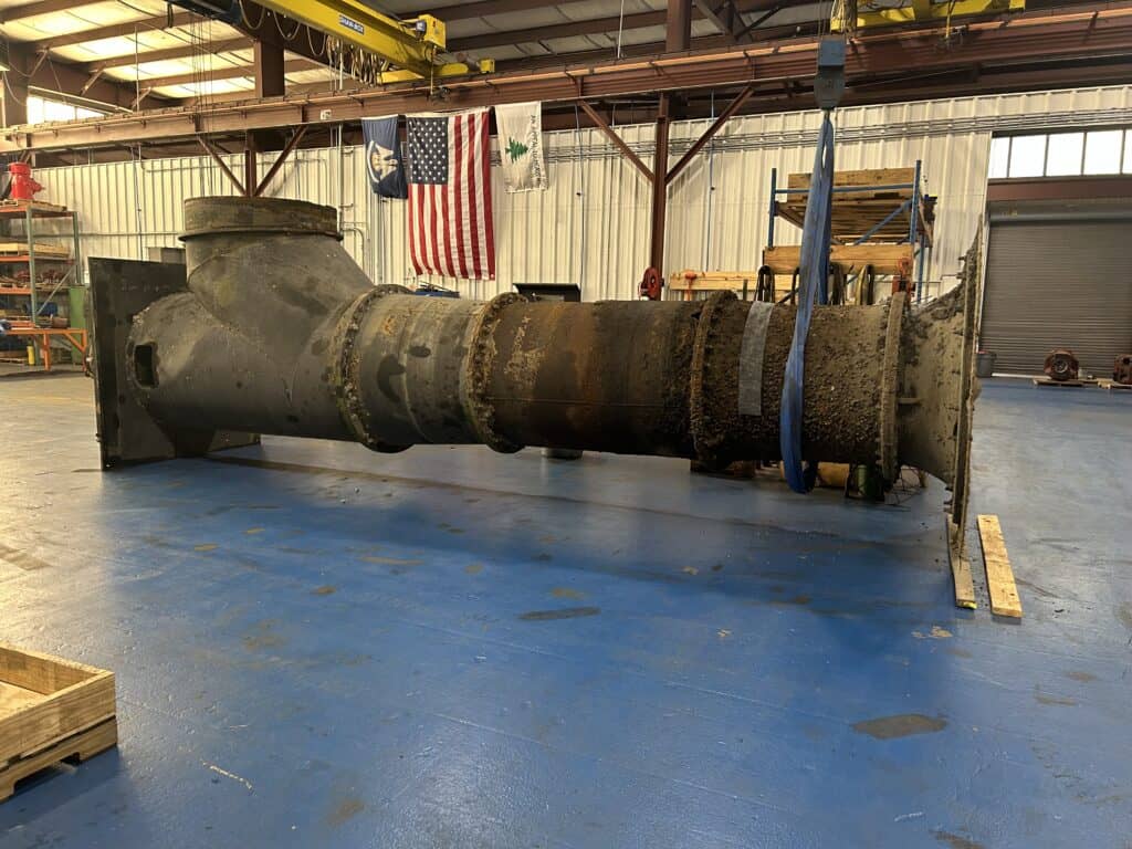 A large, rusted industrial pipe section with flanges lies on a blue floor in a warehouse with an American flag on the wall.