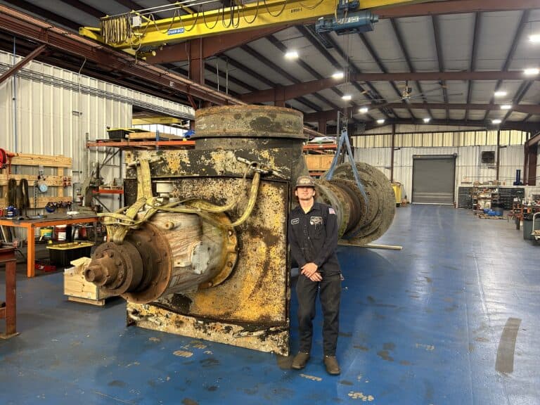 A person in work clothes stands in front of a large, rusted industrial machine inside a spacious workshop with tools and equipment.