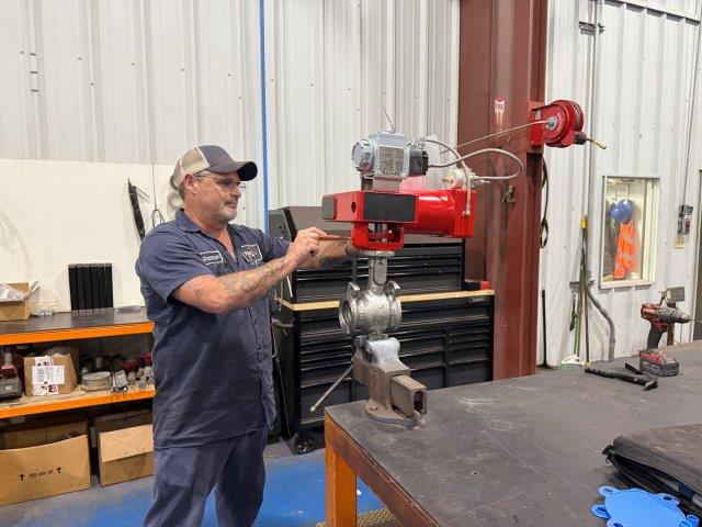 A man in work clothes operates machinery on a metal valve in an industrial workshop with tools and equipment in the background.