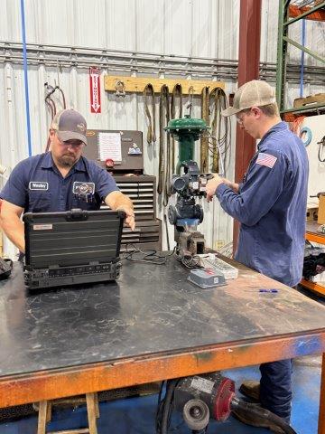 Two men in work uniforms repair industrial equipment; one uses a laptop while the other adjusts the machinery on a workbench.