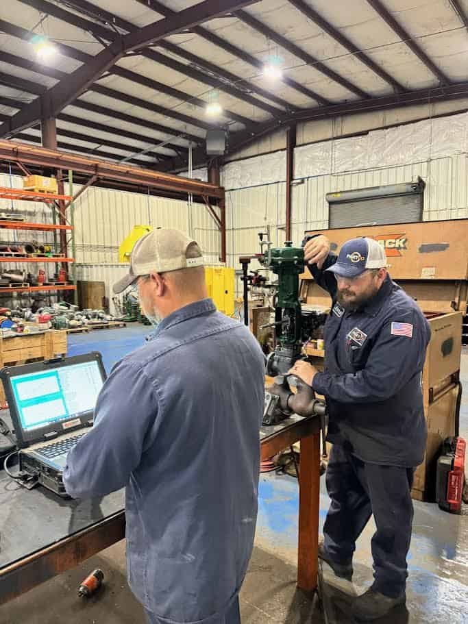 Two men in work uniforms check equipment in an industrial workshop; one uses a laptop while the other adjusts machinery.