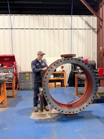 A person in work clothes stands next to a large industrial valve inside a workshop with tools and equipment in the background.