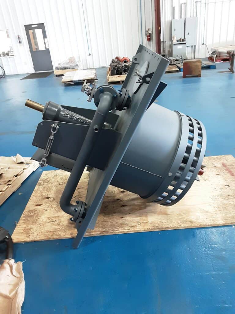 A large industrial mechanical device with metal tubing and a cylindrical base rests on a wooden board inside a warehouse.