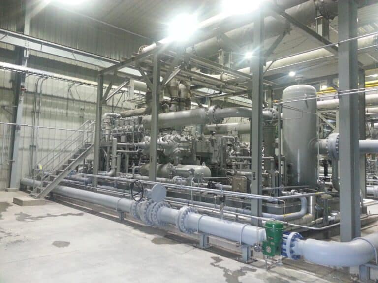 Industrial facility interior with large metal pipes, compressors, valves, tanks, and structural supports; lit by overhead lights.
