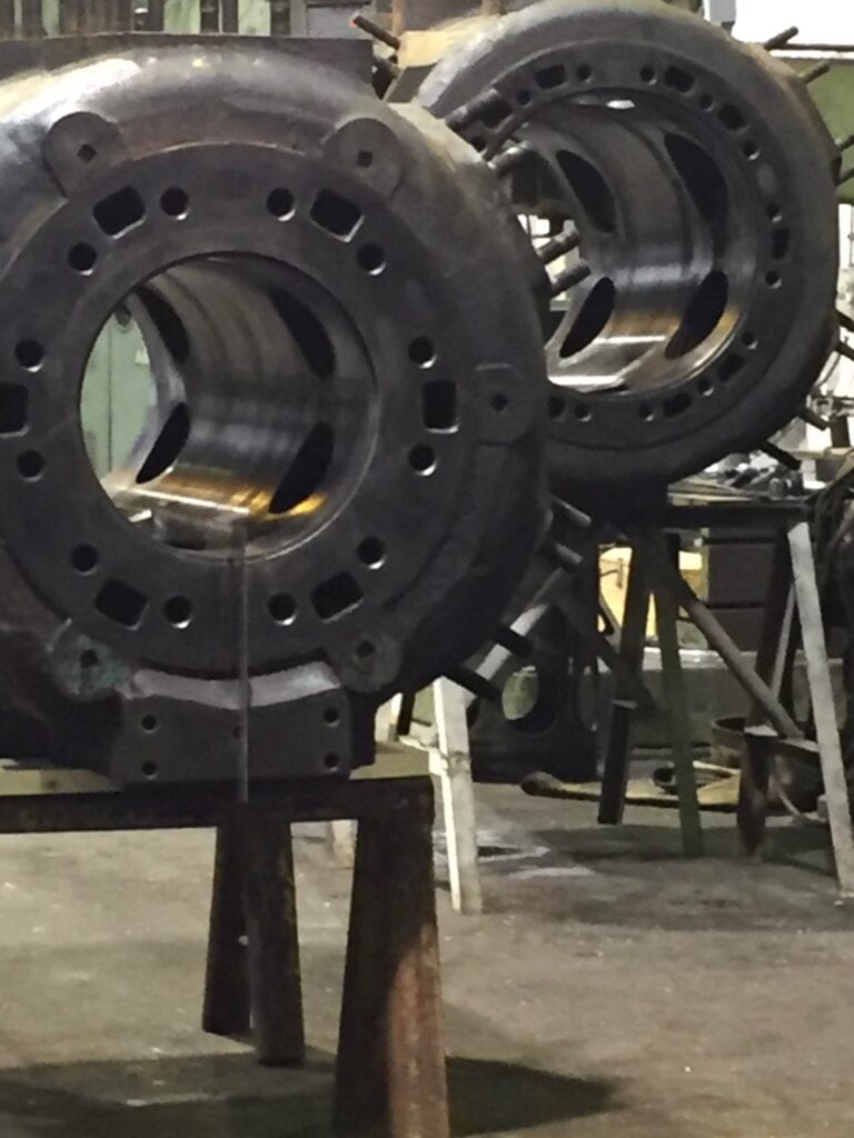 Two large cylindrical metal machine parts with circular openings are positioned on metal stands in an industrial workshop.