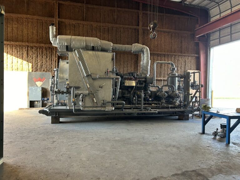 Large industrial machine with pipes and insulated ducts sits inside a spacious warehouse with a concrete floor and open bay door.