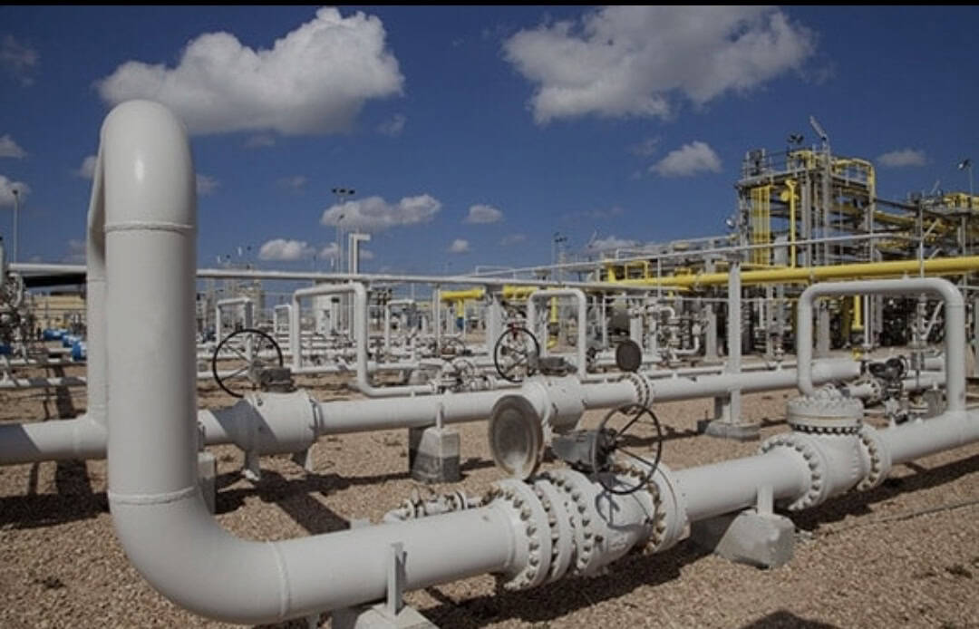 Large white industrial pipelines and valves at a gas processing facility under a blue sky with scattered clouds.