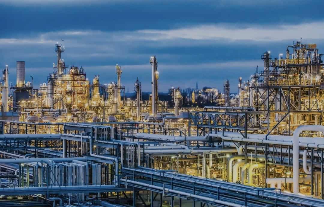 Large industrial refinery complex with numerous pipes, towers, and bright lights under a cloudy evening sky.